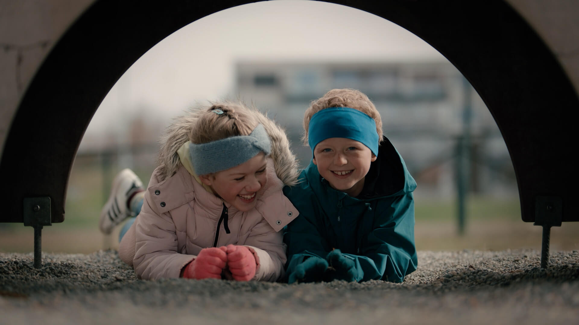 To barn ligger i grus på en lekeplass og ler sammen gjennom en buet metallkonstruksjon – et visuelt eksempel på bruk av lavt kameraperspektiv og naturlig lys i filmproduksjon for å fange nærhet og ekte øyeblikk.