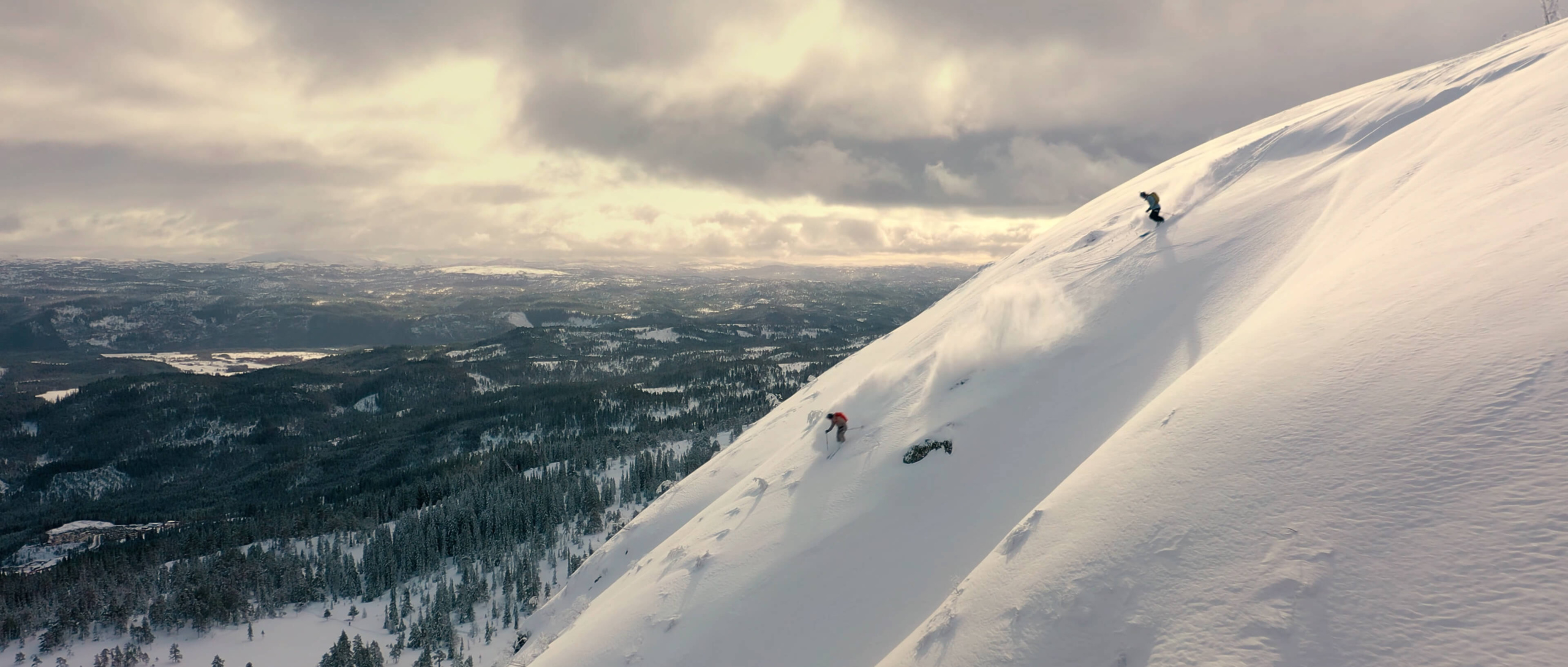 To frikjørere står på ski ned en bratt, snødekt fjellside med majestetisk utsikt over et vinterlandskap – et dramatisk dronebilde som viser hvordan filmproduksjon kan fange både action og naturskjønnhet fra luften.